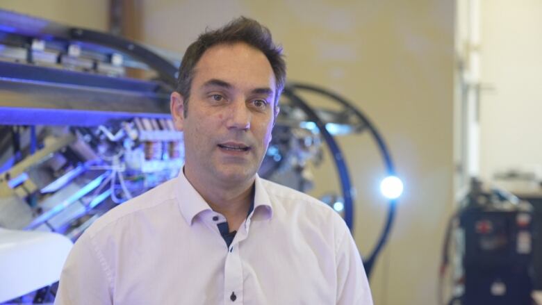 A man in a white shirt stands in a lab. It's a head and shoulders shot. He is speaking with someone just off camera