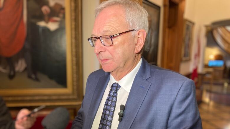 A man with grey hair and glasses addresses microphones in a hallyway.