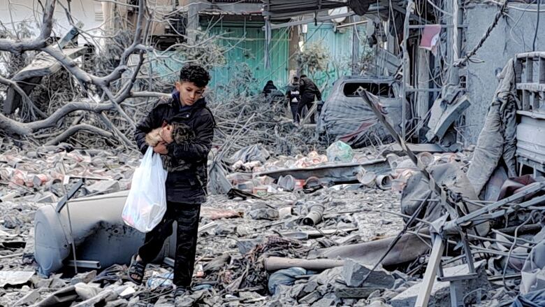 A young boy carries a cat in his arms and a plastic bag as he walks over top of pieces of broken concrete and other debris from a bombed building. 