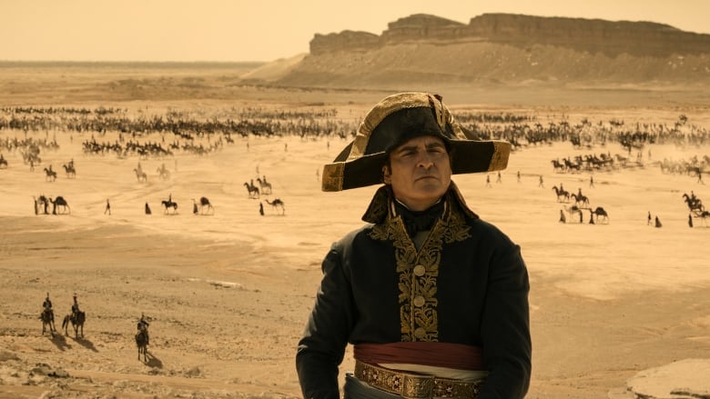 A man in an 18th century outfit looks up out of frame. Behind him is a desert, filled with cavalry. 