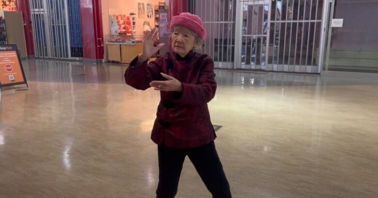 An elderly woman in a red jacket and pink hat stands in a mall with her hands in a tai chi pose. 