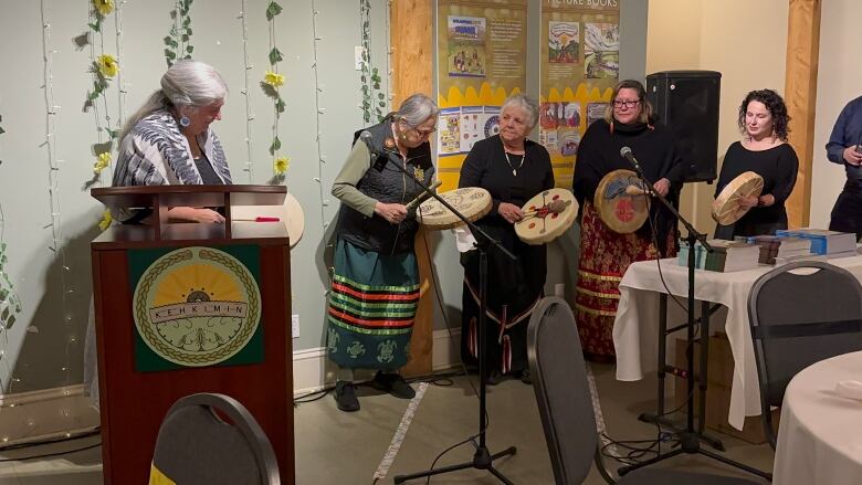 A group of women wearing long skirts and holding hand drums stand in a line in front of microphones. 