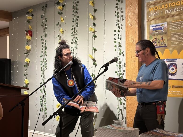 Two men, one with long hair falling down his shoulders and the other with a long braid, stand in front of two microphones holding open books.