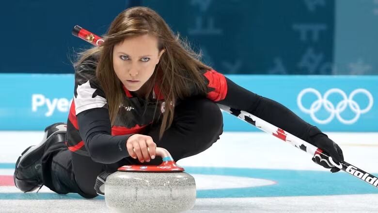 A woman prepares to throw a curling rock with one hand.. She is holding a broom in the other. 