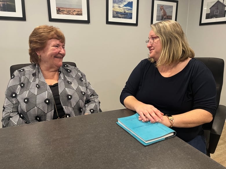 Two women look at each other and chat in a boardroom. One is clutching a blue notepad. 