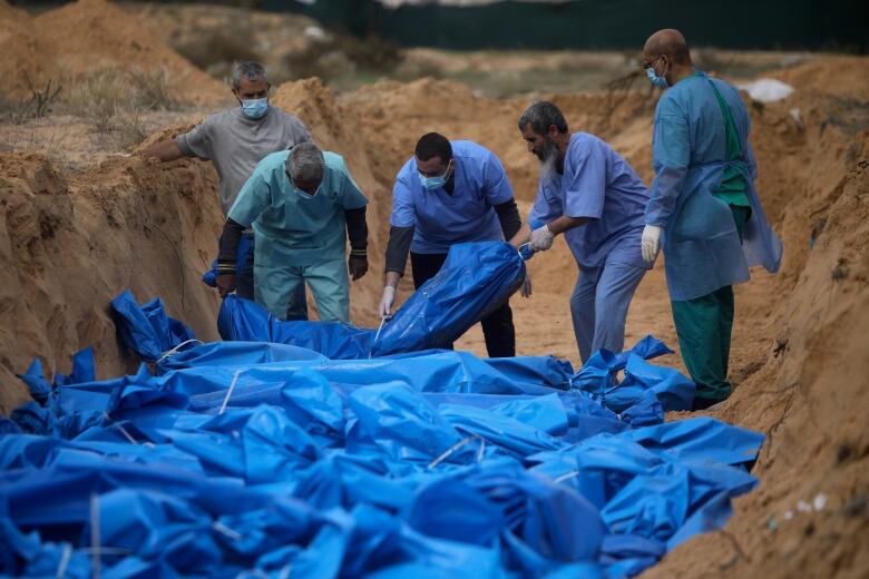 People in medical scrubs place blue plastic body bags into a mass grave. 