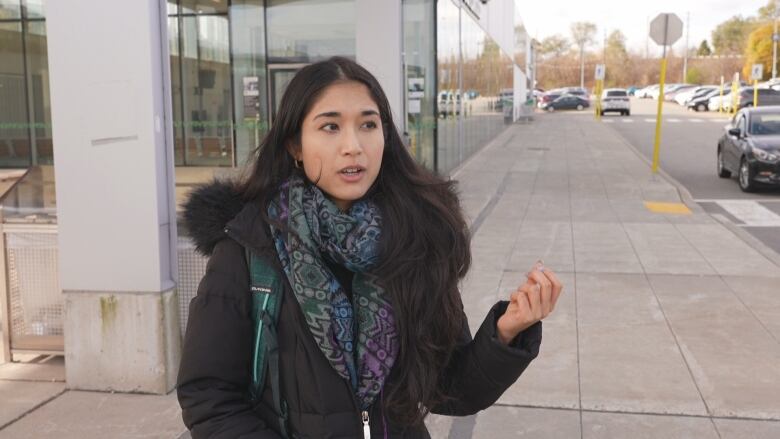 A young woman stands outside a transit station on a cold day. It is a grey day.