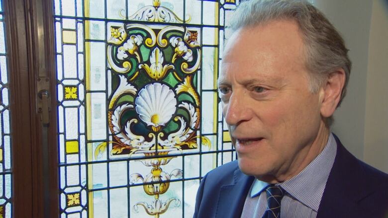 A white middle-aged man wears a blue suit, blue shirt and blue tie while standing in front of a stained-glass window talking to the camera.