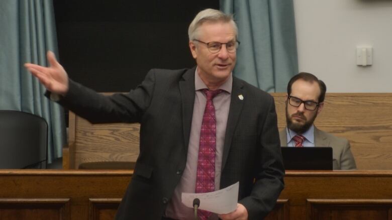 A man in a dark suit and red shirt stands up and poses questions to ministers.