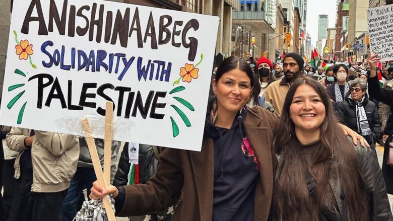 A woman at a protest holds a sign in her right hand while her left arm is draped over another woman.