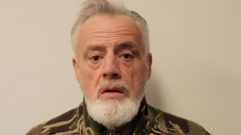 Brian Abrosimo is shown in a mug shot. He is a white man with short grey hair, a grey mustache and a white beard, wearing a camouflage jacket.