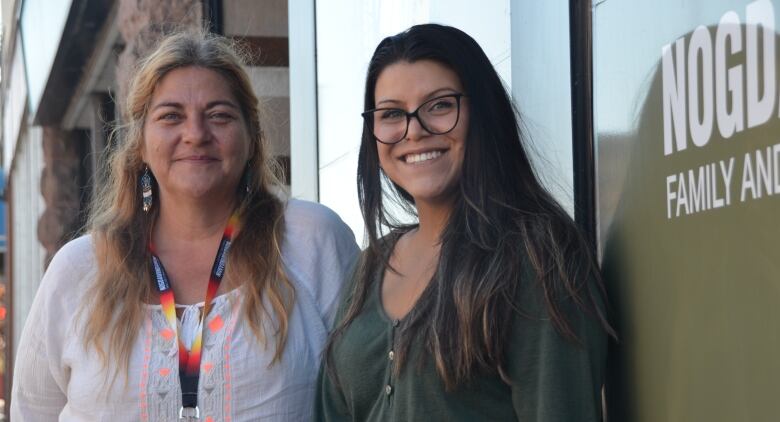 Two women smile for the camera in front of a sign on the street. 