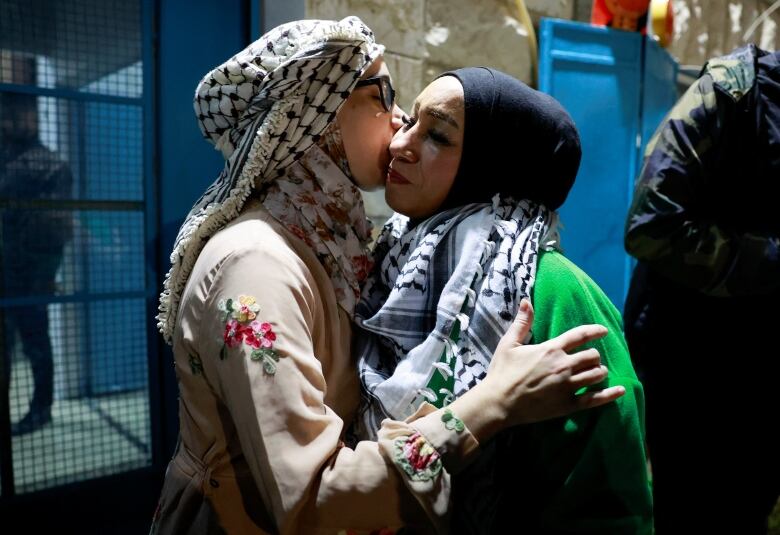 A woman wearing a white garment with flowers embroidered on the sleeve, and a black and white scarf over her hair, hugs and kisses the cheek of another woman wearing a black head-covering, a black and white scarf and a bright, green jacket. 