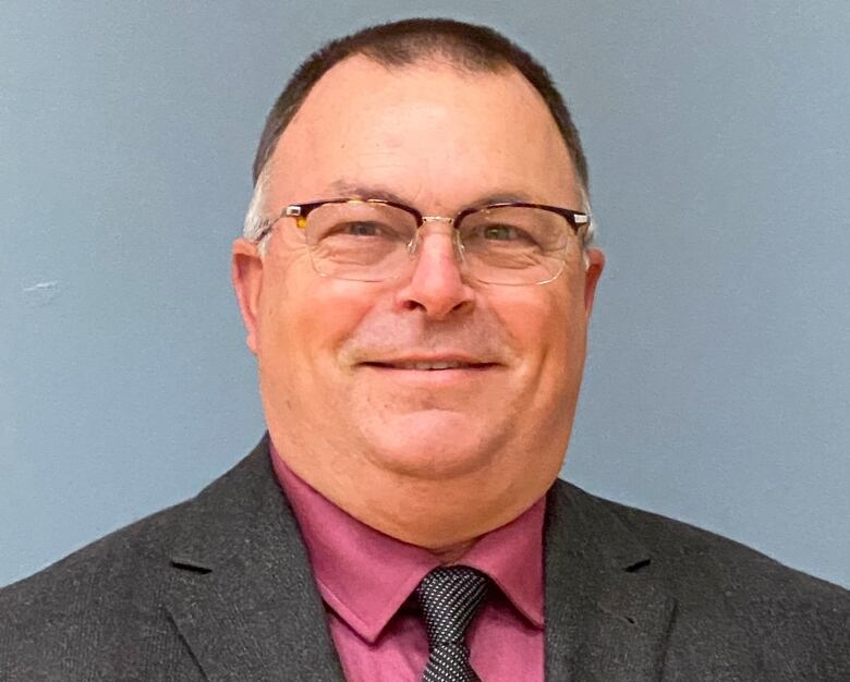 A man with glasses, a dark grey suit and magenta shirt smiles.