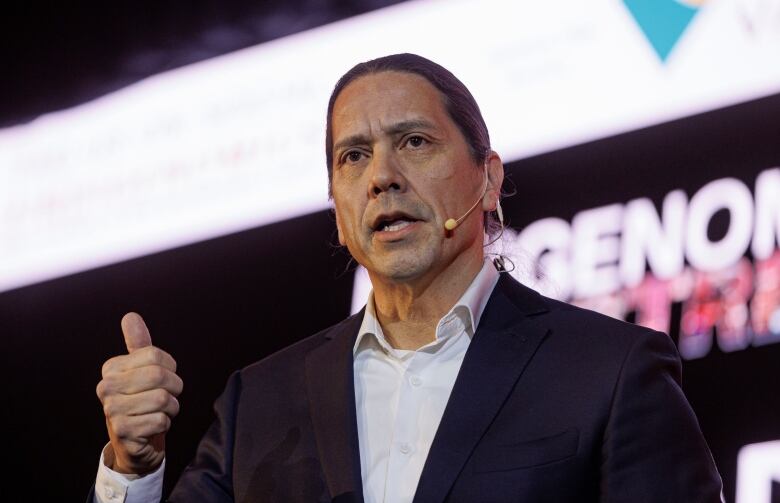 A man in a dark jacket and dress shirt without a tie speaks to a large room of people from a stage. His hair is tied back in a ponytail. 