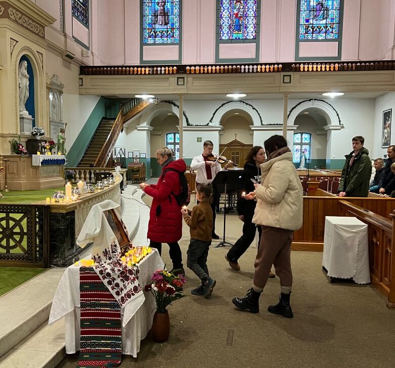 Several people can be seen walking with lit tea light candles. Dozens of candles are already resting on an altar. In the background, a young man is standing up and playing violin. 