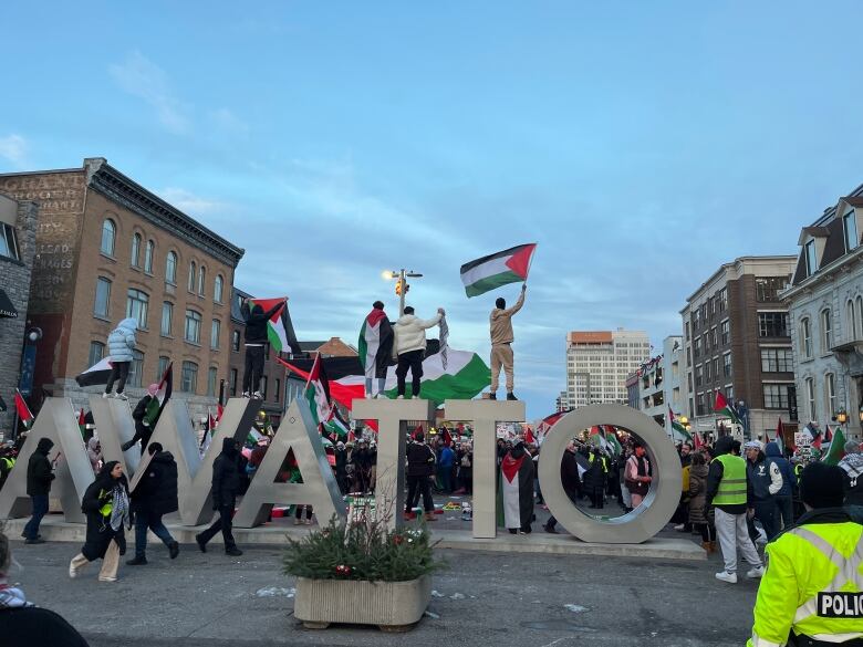 Several people wave flags while standing atop a sign reading 'Ottawa' while a large crowd is gathered in front of them.