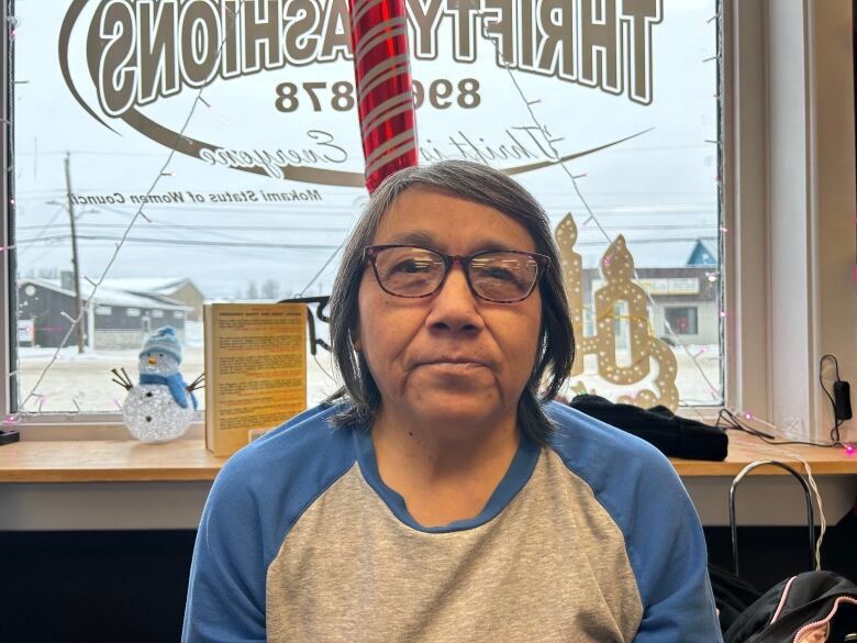 a woman wearing a gray and blue shirt and glasses sits in front of a window