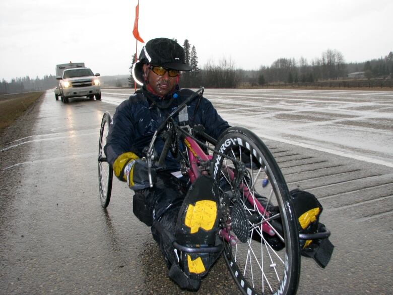 A man on a special bike designed for handcycling, peddling along a paved road in the rain. There's a truck and trailer following him. 