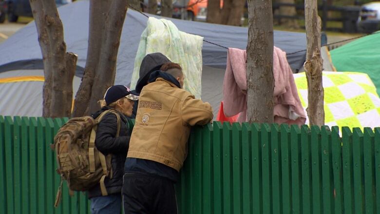 A group of people stand near a green fence with tents in the background. 