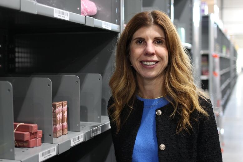 A woman with a black jacket, blue top and medium-length brown hair is pictured in front of a warehouse shelf stocked with makeup. 