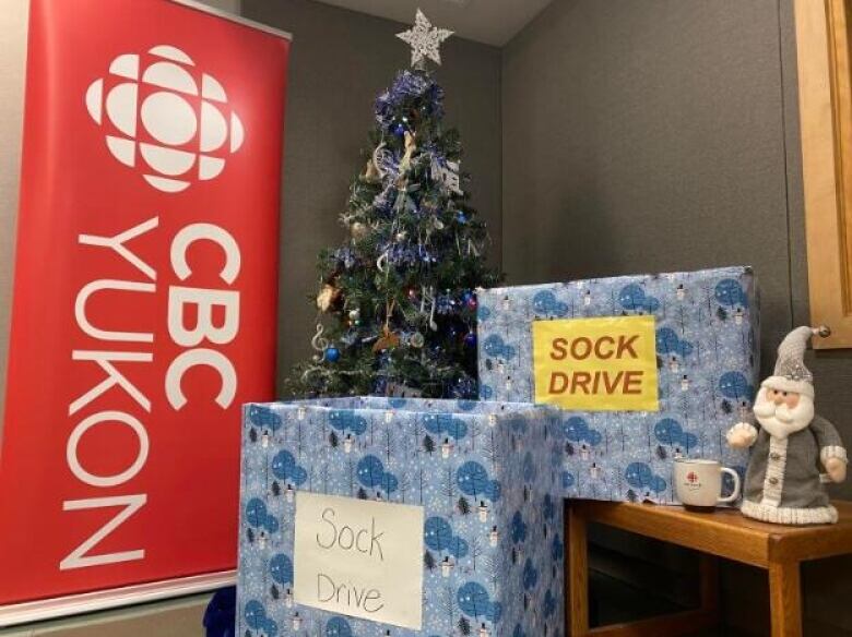 Two presents sit before a Christmas tree that stands beside a CBC Yukon pop up banner.