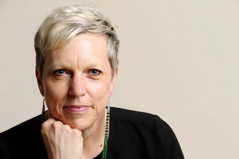 A woman wearing a black shirt and long earrings poses in front of a beige backdrop.