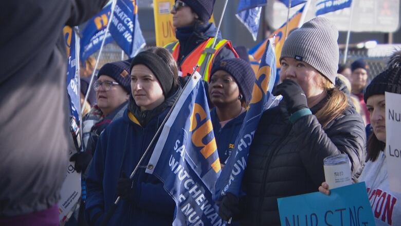 A group of women look pensively ahead as they hold blue and yellow flags reading Saskatchewan Union of Nurses. 