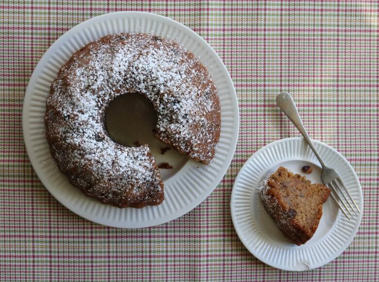 On the right, a slice of cake sits on a plate with a fork. On the left, there's a ring cake covered in icing sugar with a piece missing out of it.