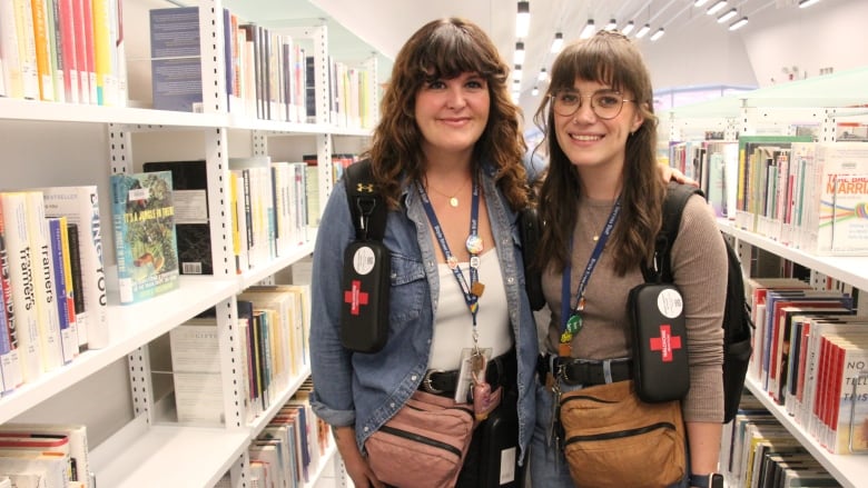 Two people pose together. Both are wearing backpacks and fannypacks. Each has a naloxone kit dangling from their backpack strap.