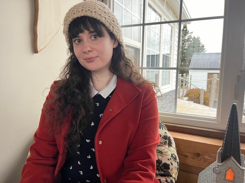 A woman with black hair and bangs wears a red winter coat and white knitted hat as she looks towards the camera. 