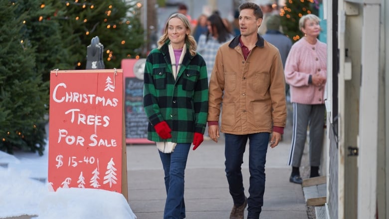 A man and woman walk down a festive street. There are Christmas trees in the background, along with a sign reading 