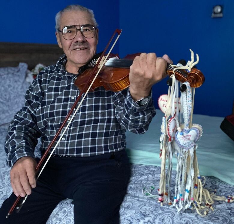 Cree elder sits on a bed with fiddle with moosehide embroidered hearts hanging from the neck.