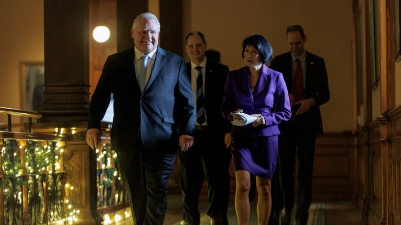 Doug Ford and Olivia Chow walk down an ornate, wood-paneled hallway with Christmas lights to their left. 