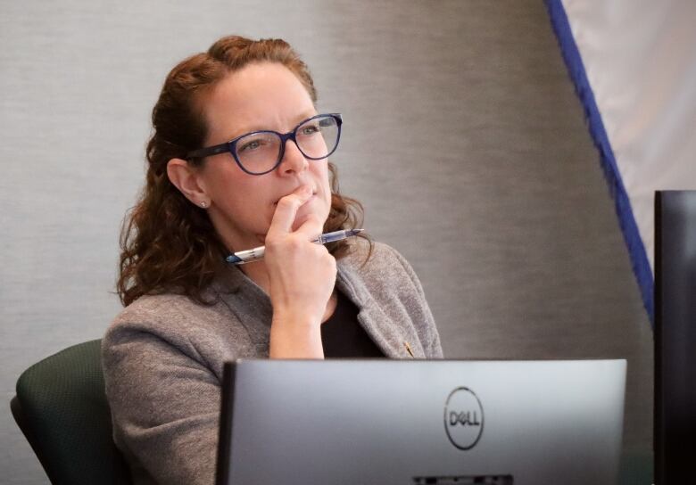 A woman in a grey jacket with red hair and blue glasses appears to be in thought, holding a pen and covering her mouth with her finger.