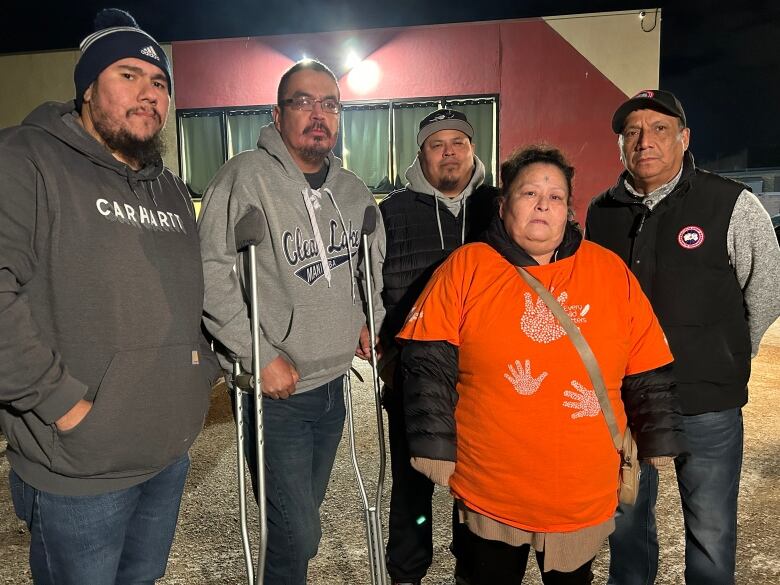 A woman wearing an orange shirt with handprints on it stands posing for a photo in front of four men in a parking lot at night.