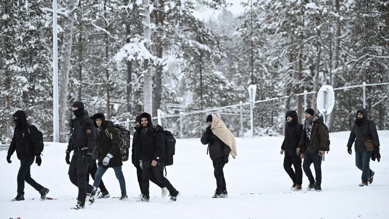 A group of men, in winter jackets with bags on their back, walk on the snowy ground with snow-covered trees in the background