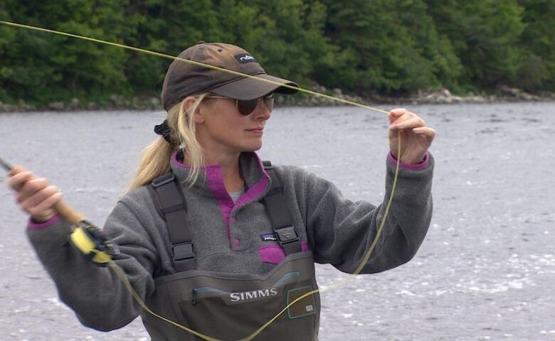 A woman wearing a cap and sunglasses raises her arms to cast a fishing line on a river. 