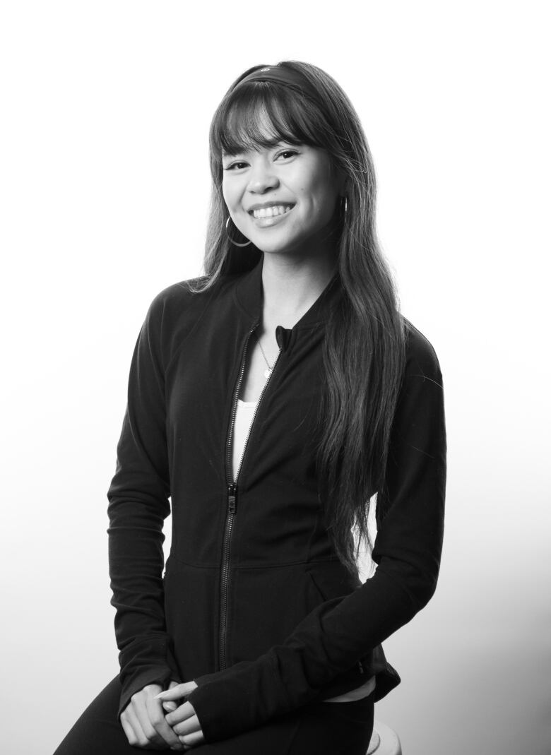 Black and white portrait of a young woman with long dark hair smiling. She is wearing a zip up jacket and clasps her hands at her lap. 