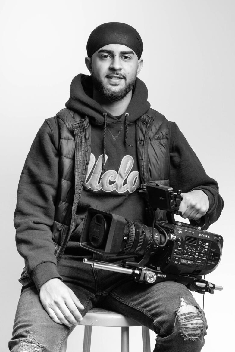 Black and white portrait of a man with light beard sitting on a stool. He holds a video camera on his lap. He is wearing a beanie, head covering, a hoodie with a UCLA logo and a puffer vest over top. He has ripped jeans.