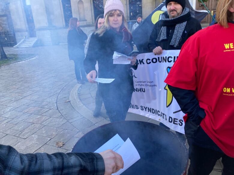 Woman with shoulder length blonde hair wearing toque and winter coat approaches barrel with piece of paper to burn.