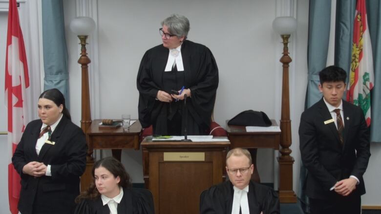 A woman in a long black robe and white tie standing while speaking to another member of the House.