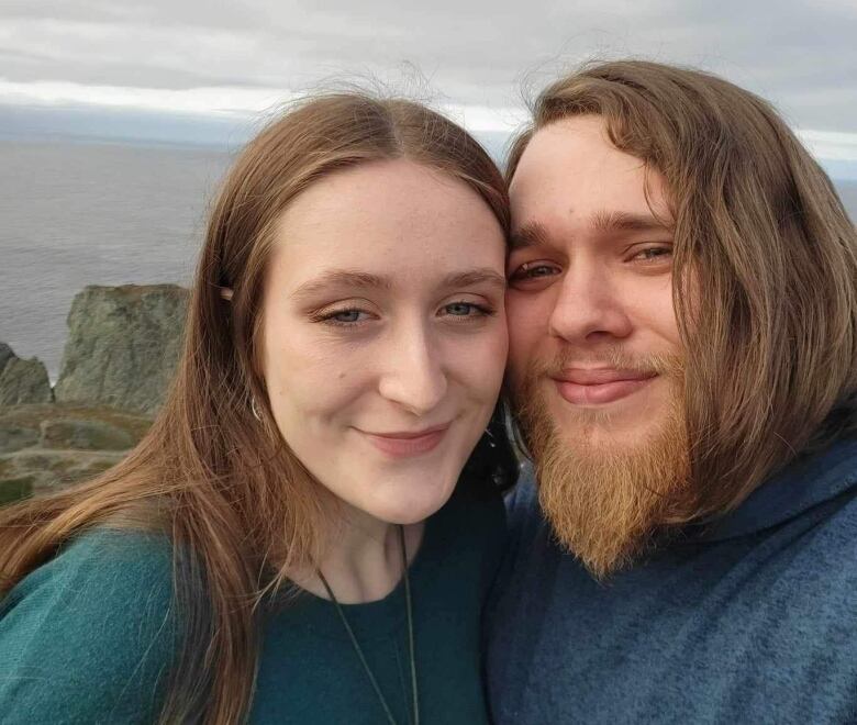 A man with a long beard holds his head close to a woman with light brown hair. They are both smiling. The ocean is in the background. 