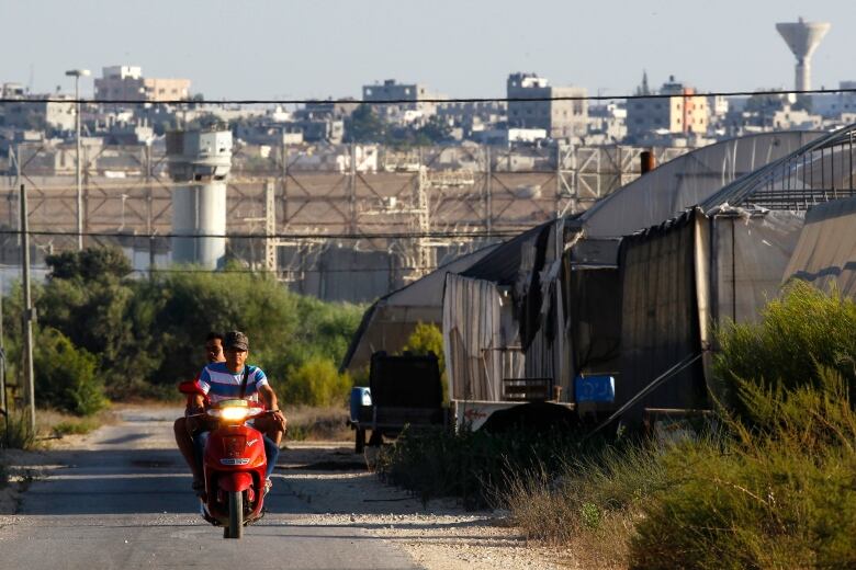 Two people ride a red scooter through a farming community.