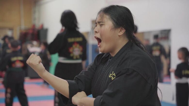 A young woman with dark hair in a pony tail yells during a martial arts class. Her hands are up towards her face and her eyes are closed. She wears a black martial arts top. 