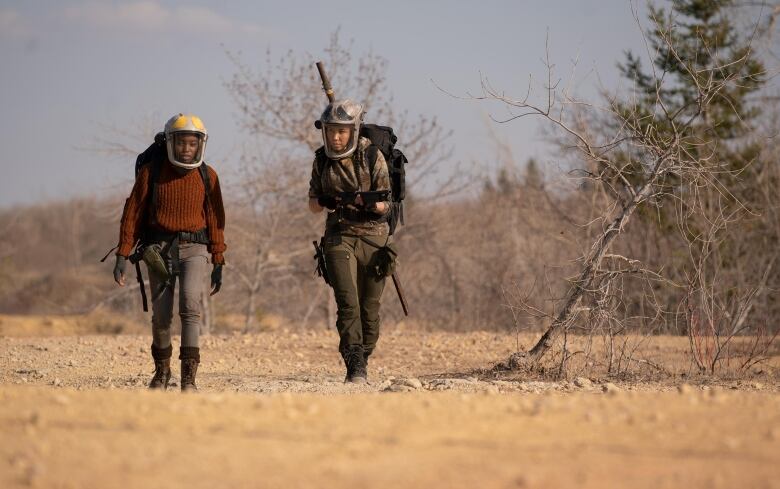 A still from a movie shows two young women in helmets with face shields walking through a dusty landscape.