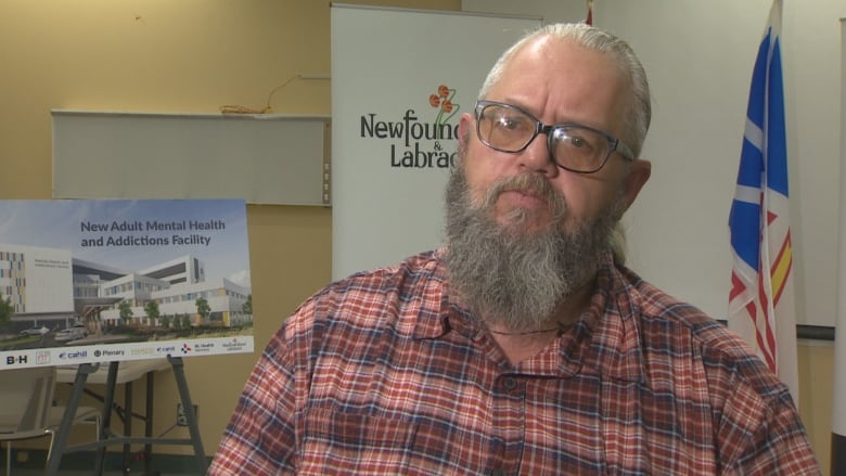 A man with a long, grey beard wearing a red flannel stands in front of a banner with the Newfoundland and Labrador logo on it.