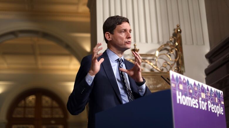 A tall white man speaks in front of a purple podium marked 'Homes for People' in an opulent hall.