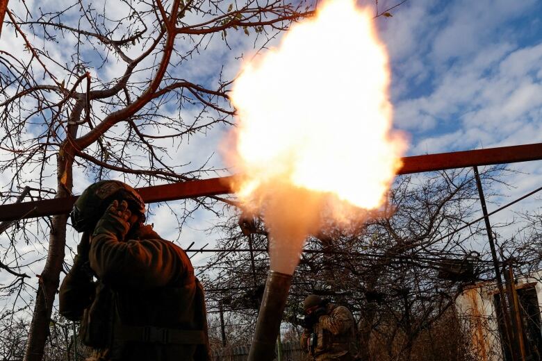 A soldier in a helmet holds his hands up to his ears as a large ball of fire and smoke escapes from a weapon.