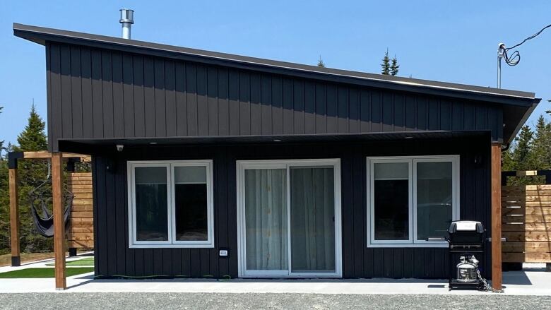 a small black building with a barbeque on the porch and fence surrounding the backyard. 